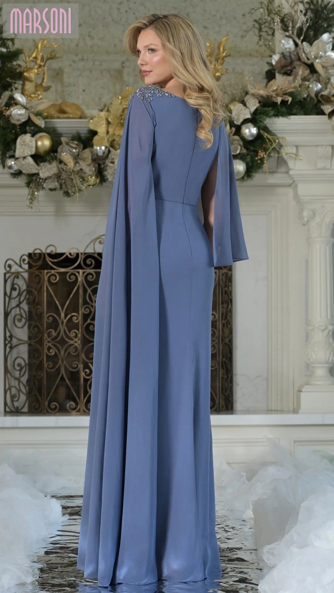 A woman in a long blue Marsoni MV1362 formal dress with cape sleeves stands indoors by a fireplace adorned with floral and gold accents.